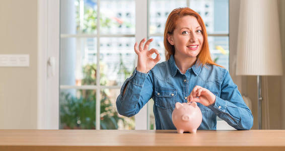 红头发的女人存钱存钱罐在家里做 ok 标志与手指, 优秀的符号