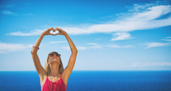 妇女在太阳镜与举起的手显示心脏标志, 当站立在海滨时