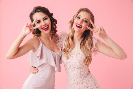 两个滑稽的女人在礼服一起玩, 同时显示和平手势, 看着在粉红色背景的相机