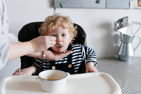 一个亲切, 关心的母亲哺养她的卷曲的学步男孩与一勺可口的汤, 家庭晚餐