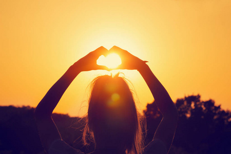 在橙色火热日落时举起手显示心脏标志的妇女的后面看法