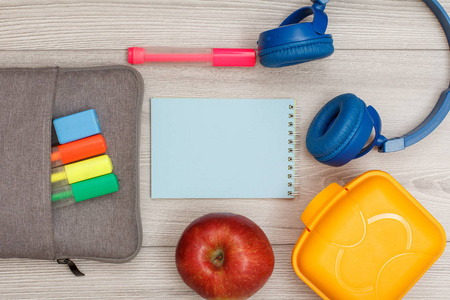 袋铅笔盒与彩色毡笔和标记, 苹果, 蓝色笔记本, 耳机和午餐盒上灰色的木质背景。具有复制空间的顶部视图。回到学校的概念。学校用品