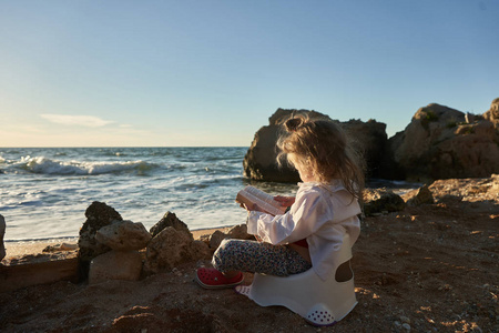 美丽的小女孩在沙滩上看书。她坐在旁边的锅, 看着离开与微笑在书中