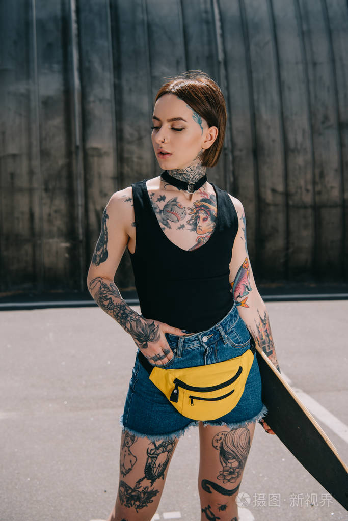 年轻迷人的纹身女孩手在口袋里拿着滑板在街上