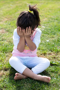 小女孩独自坐在草地上, 用双手捂住脸。