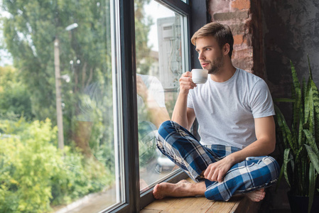 严肃的年轻人坐在窗台和喝咖啡在早晨时间在家