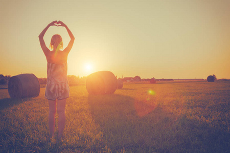 站在草地上, 在日落的干草堆的妇女与举手显示心脏标志后视图