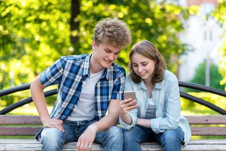 年轻夫妇在夏天一个公园室外。坐在城市的长凳上。愉快地微笑。手持智能手机的手在互联网上进行购买。露营。幸福家庭的概念