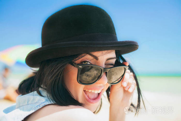 微笑着美丽的年轻女子，在黑色的帽子和复古太阳镜在海滩上的肖像