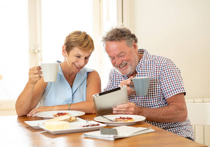快乐迷人的资深夫妇使用平板电脑上网冲浪, 同时在家里吃早饭和喝咖啡