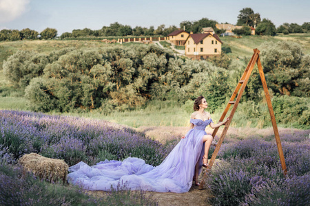 美丽的年轻女子穿着长紫色的礼服站在木制梯子上的薰衣草领域与房屋背景, 全长