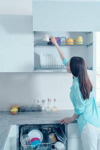 后视图的一个女人在绿松石衬衫把干净的眼镜搁在洗碗机的架子上