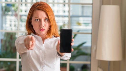 红发女子在家里使用智能手机指向相机和你, 手势, 正面和自信的手势从前面