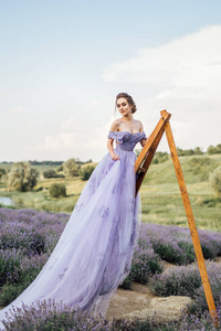 美丽的年轻女子穿着长紫色的礼服站在木梯上的薰衣草领域, 全长