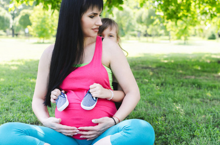 女孩抱着婴儿鞋, 拥抱她怀孕的妈妈, 在户外父亲亲吻他可爱的小宝宝在