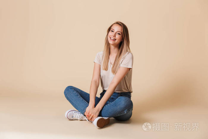 穿着休闲服装的漂亮女人的形象 smiing 坐在地板上, 腿交叉隔着米色