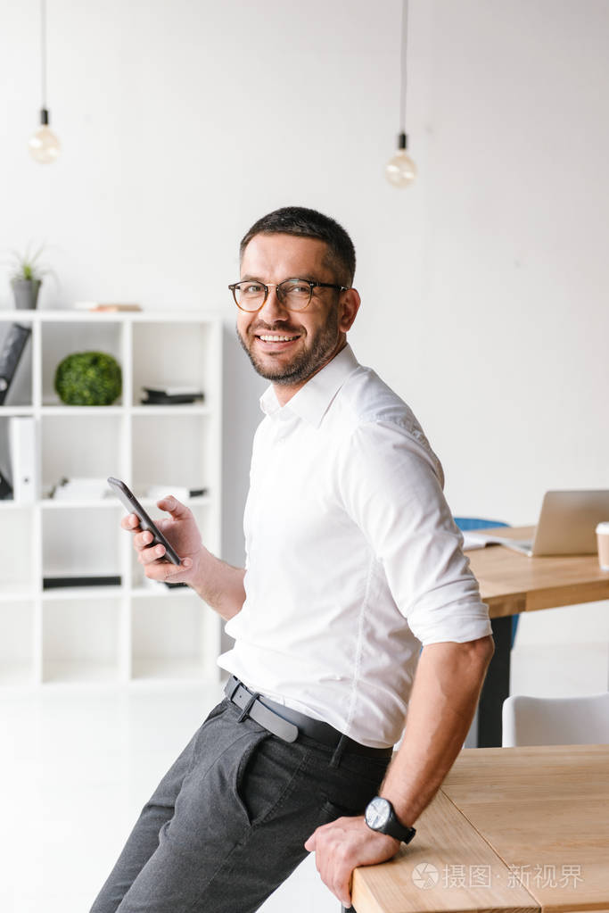 有吸引力的欧洲男人的肖像坐在桌子上的办公室和看着你, 而持有智能