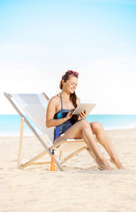 快乐的现代妇女在沙滩泳装在海滩上使用平板电脑, 而坐在沙滩椅