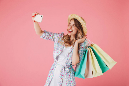 一个快乐的年轻女子的肖像在夏季礼服和草帽采取自拍, 而持有购物袋孤立的粉红色背景
