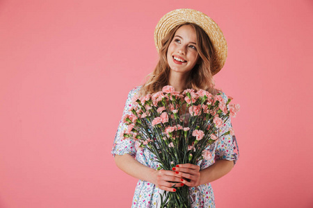 一个欢快的年轻女子在夏季礼服和草帽捧着康乃馨花束和看着远离粉红色背景的照片