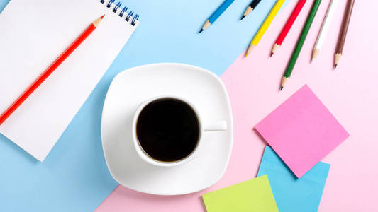 白杯, 有黑色的咖啡碟, 彩色铅笔, 贴纸, 笔记本。粉红色和蓝色背景。平躺。经营理念。柔和色调