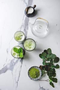 旳绿茶冰拿铁或鸡尾酒在三不同的杯子与冰块, 旳粉和牛奶罐在白色大理石桌, 被绿色的分支装饰。背景灰色墙壁