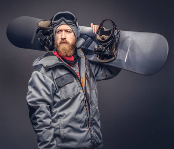 野蛮的红发滑雪板在一顶冬天的帽子和防护眼镜, 穿着滑雪板外套摆在一个工作室的滑雪板大衣满胡子。在灰色背景上隔离