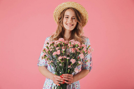 一个微笑的年轻女子在夏季礼服和草帽持有康乃馨花束隔离粉红色背景的肖像