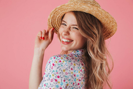 关闭在夏季礼服和草帽摆出一个笑的年轻女子的肖像和看着在粉红色背景隔离相机