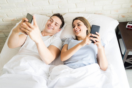 微笑的夫妇使用智能手机, 而在床上放松在家中
