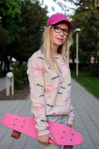 在公园的粉红色的帽子和滑板金发女孩的肖像。女孩在外面的眼镜在街上。童年, 夏天, 假期, 运动