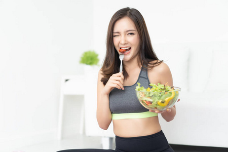 年轻的亚洲妇女正在休息和吃健康的沙拉后, 锻炼和眨眼她的眼睛, 微笑着看着在客厅的相机。健身和健康的生活方式概念。绿色健康食品概