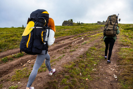 旅行生活方式和生存概念后视图。两个徒步旅行的妇女带着背包沿着小路走到山上, 在一个温暖的夏日里长途跋涉, 后面的景色