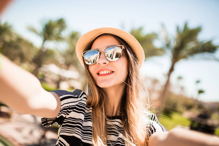 微笑在帽子和太阳镜以夏季度假村棕榈树背景拍照手机从手中的女人。暑假的时候