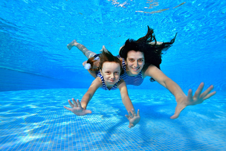 快乐的女儿与她的母亲潜水水下到池底的条纹泳装, 看着相机和微笑的蓝色背景。水下摄影。景观定位