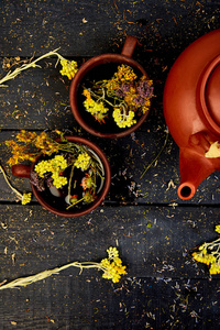 两杯草药茶tutsan, 艾树, 牛至, 蜡菊, 薰衣草附近棕色茶壶在黑暗的木质背景。凉茶。干草药和花卉, 草药。平躺