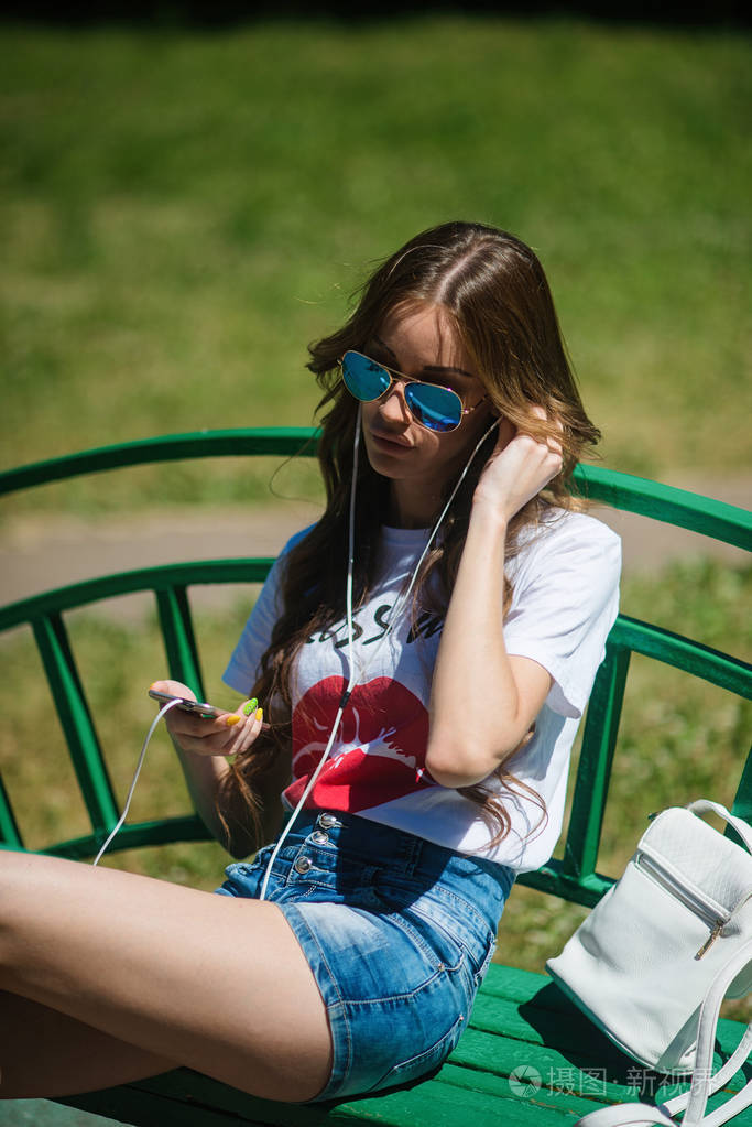 年轻美丽的女孩听音乐的耳机, 坐在长椅上的公园