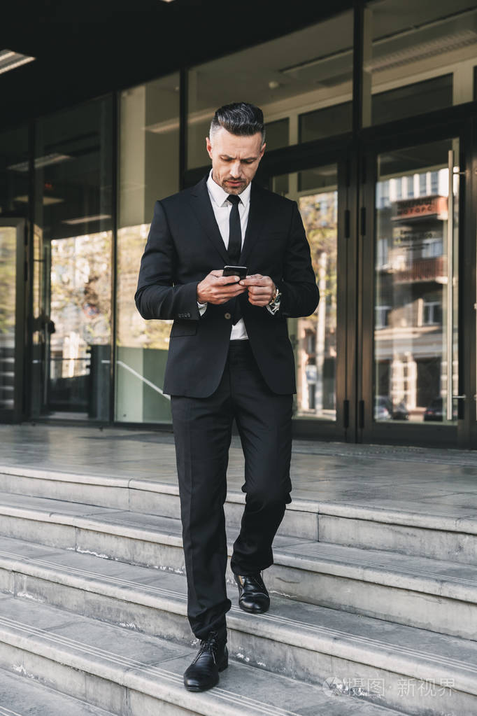 一个成功的年轻商人的肖像穿着西装走下楼梯外的玻璃建筑和使用手机