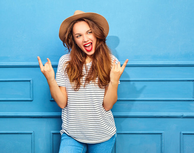 年轻时尚的女孩在休闲夏天的衣服和棕色的帽子, 红色的嘴唇, 在蓝色的墙壁上的造型。显示摇滚标志