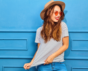 年轻时尚的女孩在休闲夏天的衣服和棕色的帽子, 红色的嘴唇, 在蓝色的墙壁上摆出的模型