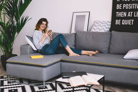 快乐的年轻女子在家里享受自由的时间躺在舒适时尚的沙发上, 看有趣的视频在社交网络上的智能手机设备。积极的博客在聊天中的留言笑