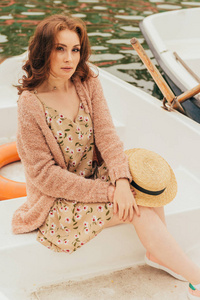 那女孩坐在河上的一条白色小船上。身着花裙, 米色毛衣, 手里拿着一顶太阳帽。暑假