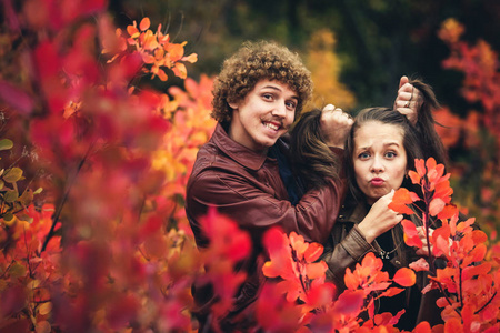 欢快的情侣是顽皮的, 并显示在红色的秋天树之间的情绪。卷曲头发的大胡子人笑着, 拿着头发, 女孩曲折的脸