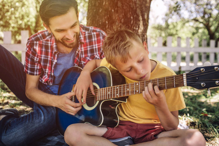 有才华的男孩正在看弦和弹吉他。他的父亲坐在他旁边, 帮助他。成人微笑