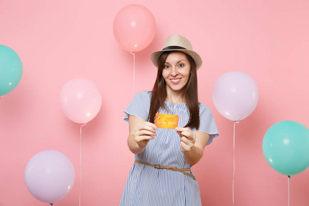 微笑迷人的年轻女子在稻草夏季帽和蓝色礼服持有信用卡在柔和的粉红色背景与彩色气球的肖像。生日派对人真挚的情感