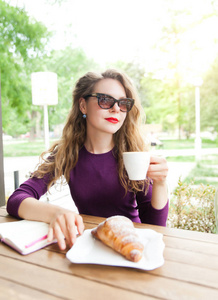 Beautifl 迷人的年轻金发女郎, 卷曲的头发喝卡布奇诺和吃牛角面包在城市咖啡馆的夏日。笔记本和电话在桌子上