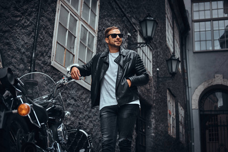 时尚的车手在太阳镜身着黑色的皮革夹克和牛仔裤靠在他的复古摩托车在旧欧洲街
