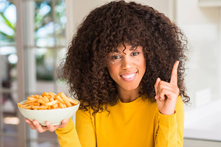 在家里拿着薯片的非洲裔美国妇女惊讶于一个想法或问题手指与快乐的脸, 第一