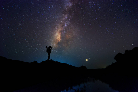 银河有星星和夜空与背包的剪影。手持相机和三脚架站在山上