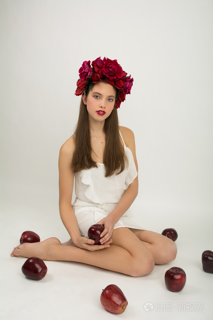 年轻漂亮的女孩在用花环的她头上的花和苹果在白色背景上的白色夏装